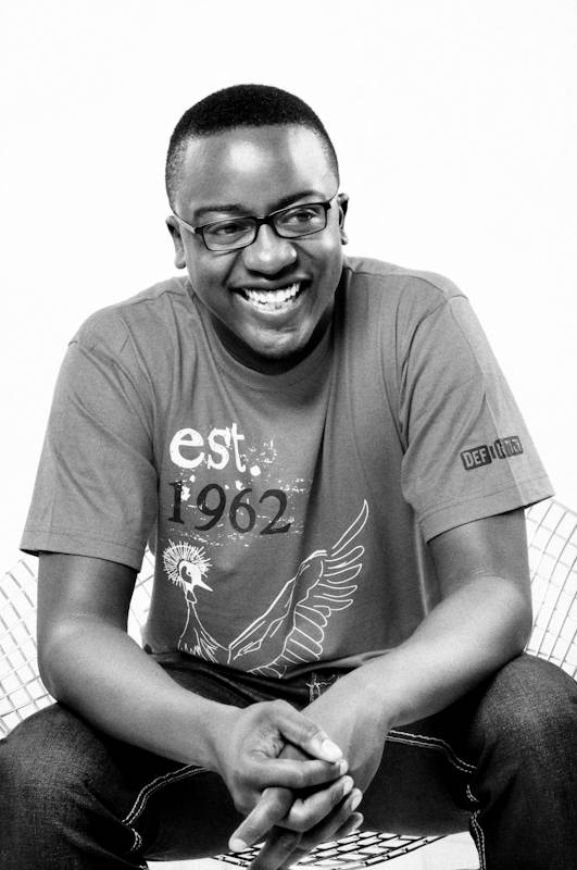 Mister Deejay, smiling, seated with hands clasped, in a black and white portrait.